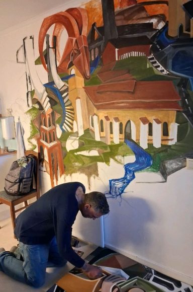 En man knäböjer framför en väggmålning med färgglada mönster och former.